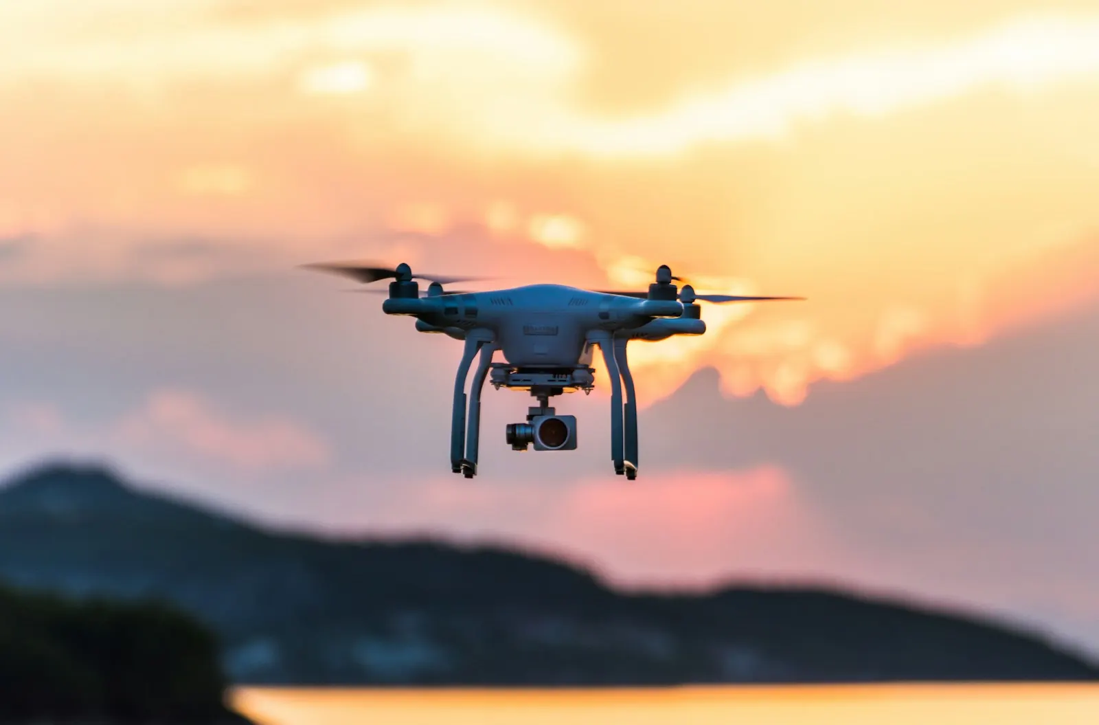 A white quadcopter drone in the air at sunset over a beach, its camera facing forward